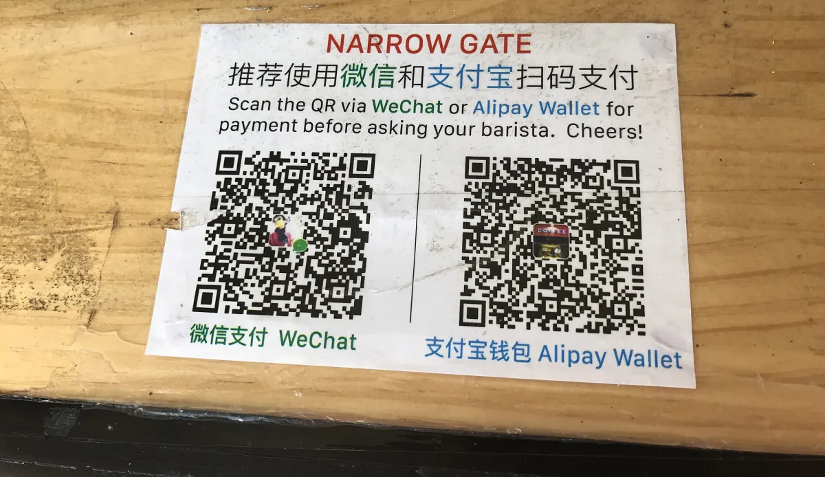 A printed café sign with separate WeChat and Alipay payment QR codes on a wooden counter